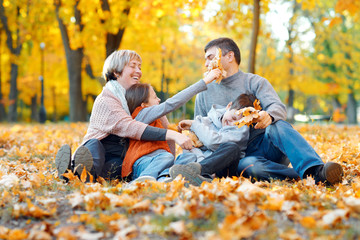 Happy family sitting on fallen leaves, playing and having fun in autumn city park. Children and parents together having a nice day. Bright sunlight and yellow leaves on trees, fall season.