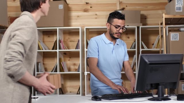 Thigh-up shot of Middle Eastern male postal service clerk, wearing blue uniform polo shirt, serving customer in office, giving away cardboard package and registering it on computer