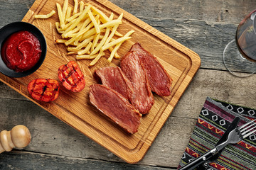 Steak with French fries and grilled cherry tomatoes on a cutting board. Classic meat meal with red wine on an ethnic tablecloth. Top view, directly above.
