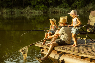 Cute little girls and their granddad are on fishing at the lake or river. Resting on pier near by water and forest in sunset time of summer day. Concept of family, recreation, childhood, nature.