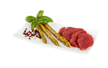 A small plate with asparagus, herbs and cured sausage on a white background.