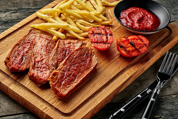 Steak with French fries, grilled cherry tomatoes, sauce and red wine on a rustic wooden table. Wholesome American lunch served on a cutting board.