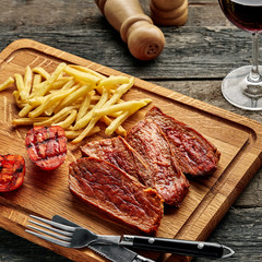 Steak with French fries and grilled cherry tomatoes on a cutting board. Classic meat meal with red wine on a wooden table with black tablecloth.