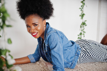Fototapeta premium Attractive african american woman with afro hair wear on skirt and jeans jacket, posed at white room on swing. Fashionable black model.