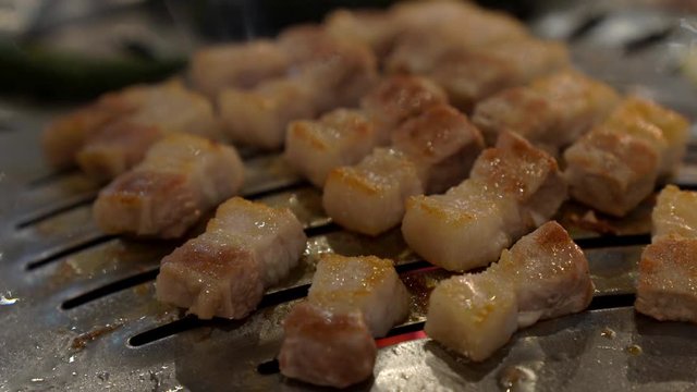 4K Close up shot of chef hand cooking korean charcoal smoked grilled pork belly meat with grilled green chilli and garlic on iron grill pan in korean restaurant. Korean BBQ, food and drink concept.