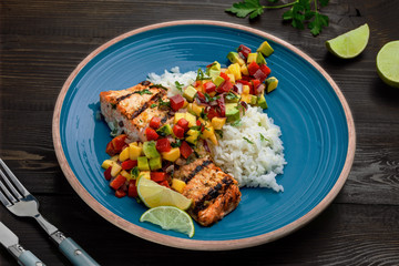 Grilled fillet of salmon with rice garnish and exotic fruit sauce made of mango, avocado, pepper and onion on a rustic wooden table.