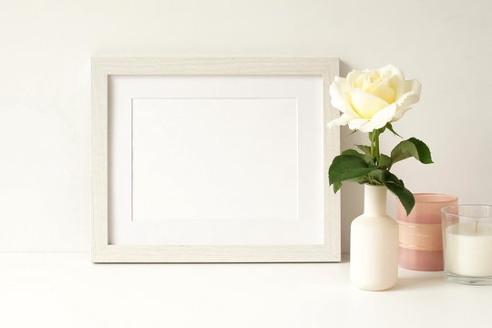White frame mock up on white wall, rose in a vase, decorative candles . Home decor close up. Copy space