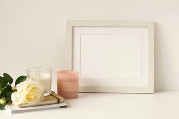 White frame mock up on white wall, rose, decorative candles . Home decor close up. Copy space