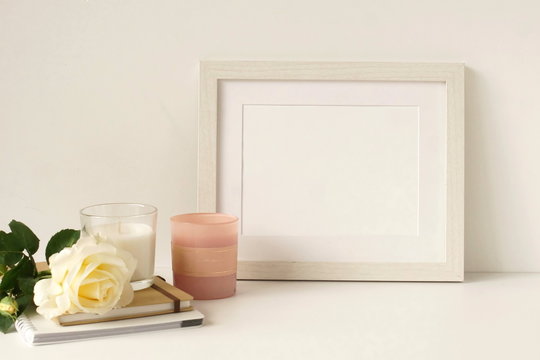 White frame mock up on white wall, rose, decorative candles . Home decor close up. Copy space