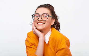 Portarit of cheerful student girl smiling broadly, wears orange sweater and round transparent eyewear. Pretty positive young woman feels joyful posing over white studio wall. People and emotions