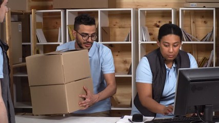 Thigh-up shot of male Arab and female Afro-American post office clerks working together, man bringing cardboard boxes to counter, woman scanning and processing them on computer, and giving to courier