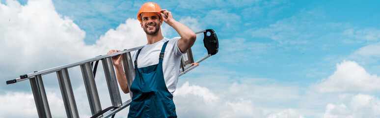panoramic shot of cheerful repairman holding ladder and smiling against blue sky with clouds © LIGHTFIELD STUDIOS