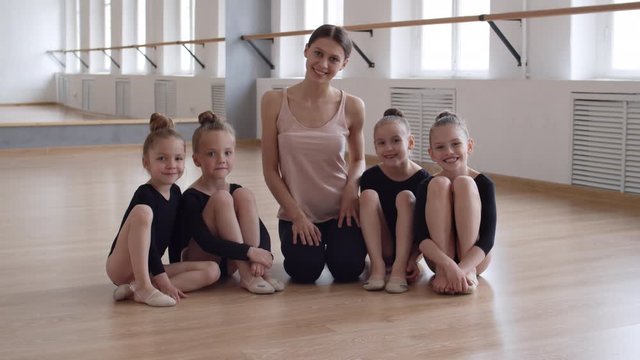 Lockdown of young female ballet teacher sitting on floor in ballet class surrounded by little Caucasian girls wearing black costumes sitting nearby. They looking at camera and smiling
