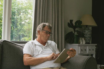 Active senior man sitting on sofa and using digital tablet in living room at comfortable home