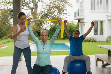 Female trainer assisting active senior couple to exercise with resistance band in the porch