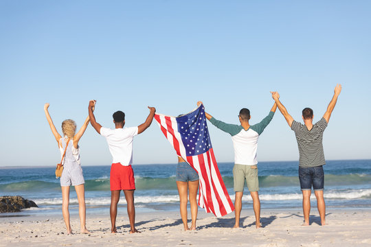 Group of friends standing together with American flag and raising hands on the beach - Powered by Adobe