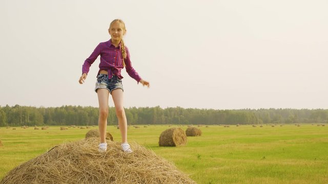 Pretty Girl Dance Haystack Mow Crop Straw Coil. Happy Caucasian Child Musical Movements Hay Bale Top. Cute Blonde Teenage Casual Clothes Summertime Harvest Farm Field Landscape Distant Wood Background