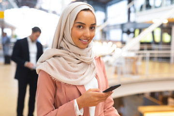 Businesswoman in hijab looking at camera while using mobile phone in a modern office