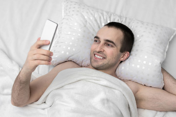 Joyful man woke up in the morning in his bedroom, man uses smart phone, top view