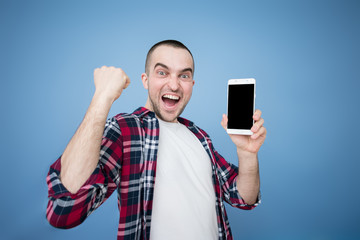 Obraz na płótnie Canvas Funny young man shows screen smartphone, screaming: Yes! Winner!, blue background, copy space