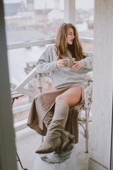 Relaxed woman with cup of coffee or tea. Stylish girl dressed in a gray sweater and silk skirt resting on the balcony.