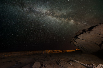 The Milky Way in the night at the rocky coast of La Mata. A big rock juts like a roof into the sky, the ground is rocky and jagged. A lantern gives some warm light.