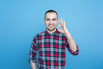 Portrait, smiling young guy shows okay, looking at camera, blue background, copy space