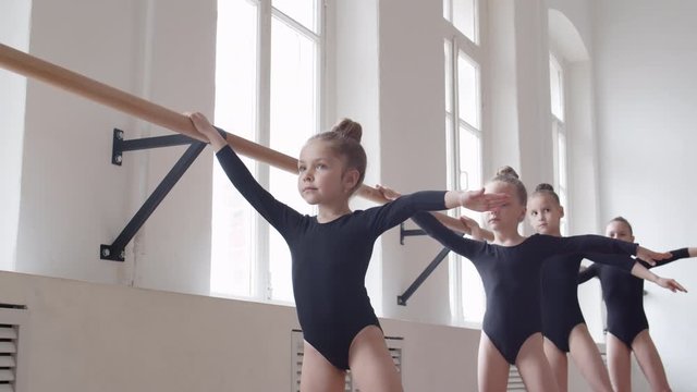 Female ballet teacher helping four little Caucasian girls wearing black costumes standing in row and practicing ballet using stick on the wall