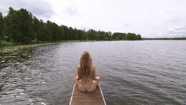 Slow gimbal meditating pan: Fit slim woman practicing yoga exercises wearing mini swimsuit bikini at lake with clouds - Yoga meditation and wellness lifestyle concept - Full sitting shot