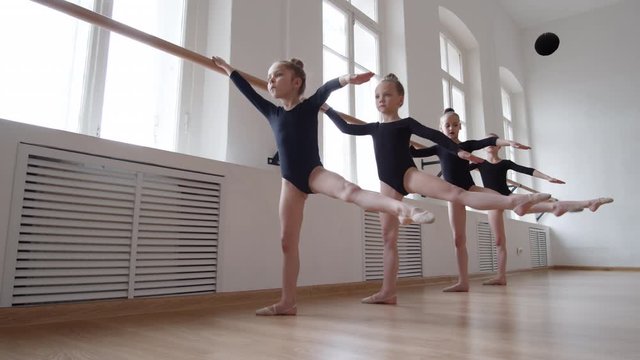 Wide shot of four little Caucasian girls wearing black costumes standing in row and practicing ballet using barre