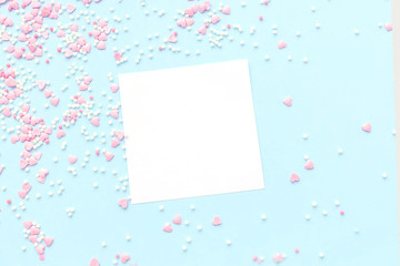 Festive romantic gentle abstract background for the design. Pink confetti in the shape of hearts and blank white sheet of paper for text  on a blue background. Top view, flat lay, copy space.