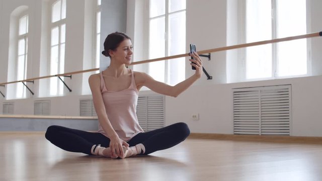 Panning of young pretty woman sitting on floor in ballet studio, stretching her legs while making selfie
