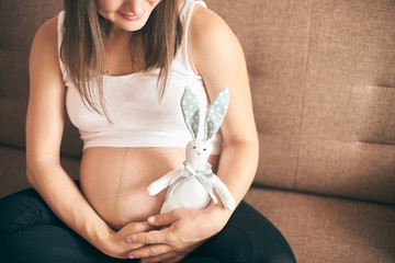 Front view of happy pregnant woman sitting on sofa at home, embracing big belly and keeping cute...
