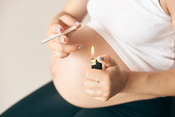 Selective focus of lighter and cigarette in hands of pregnant woman. Female in white shirt smoking during pregnancy period. Concept of threat and harmfulness.