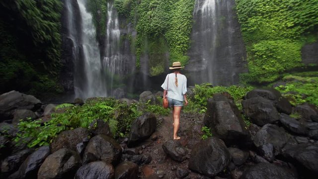 Barefooted woman in shorts and straw hat is walking through small pond on wet rocks and enjoying incredible waterfall Sekumpul (95m) in Bali Rainforest. Indonesia