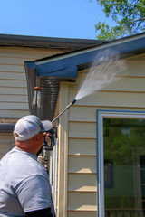 Man wearing a white baseball cap power washing the siding and roof of a beige and blue house