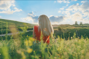 Blond business woman in a red dress resting sitting on the grass in the sunset time, view from the back