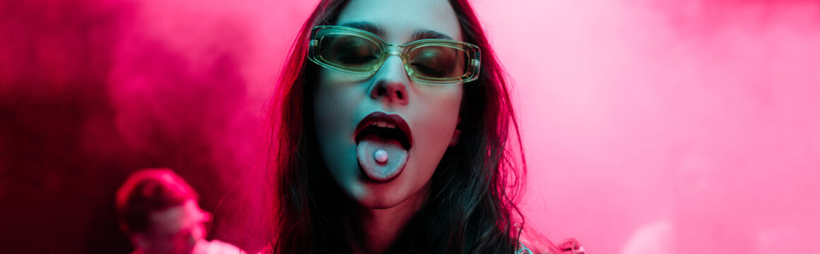 panoramic shot of beautiful girl in sunglasses with lsd on tongue in nightclub with pink smoke