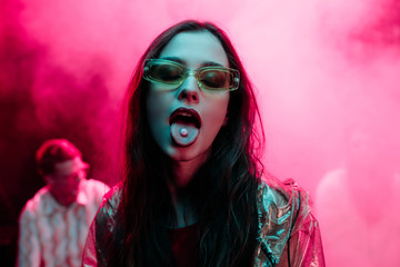 beautiful girl in sunglasses with lsd on tongue in nightclub with pink smoke - obrazy, fototapety, plakaty