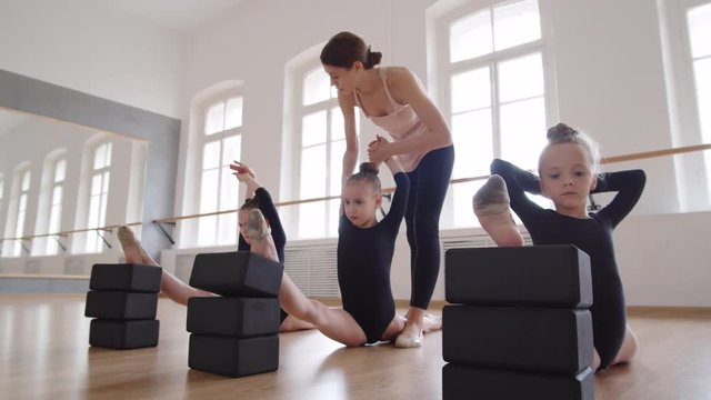 Tracking left of three little girls in black leotards sitting on floor in twine using yoga blocks with hands above their heads while young female ballet teacher going along room and helping them