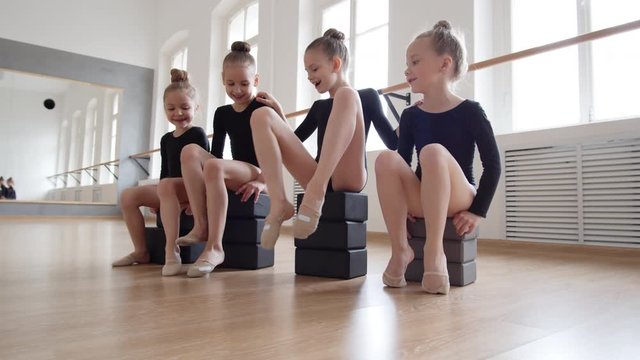 Pan shot of four little Caucasian girls wearing black costumes sitting on yoga blocks in ballet studio, having fun and laughing