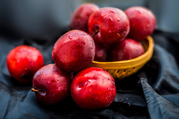 Raw organic red ripe plums in a brown-colored basket on black fabric on the wooden surface.