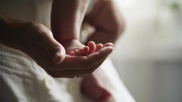 Mother with two month child on hands standing opposite window in daylight. Detail view woman gently touch little baby feet and fingers in hand. Conceptual image of maternity.