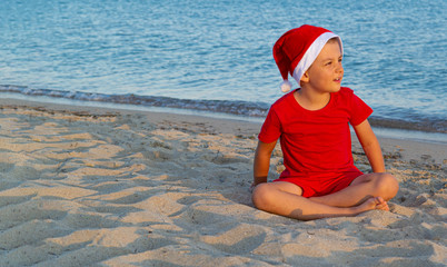 A child in a red Christmas hat playing on the sea. Santa boy. The concept of Christmas holidays at...