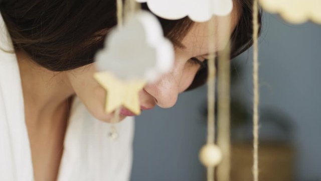 Mother standing near the cradle and bending over lying baby. Close-up view through defocused hanging toys above cot, portrait young pretty woman caring for child