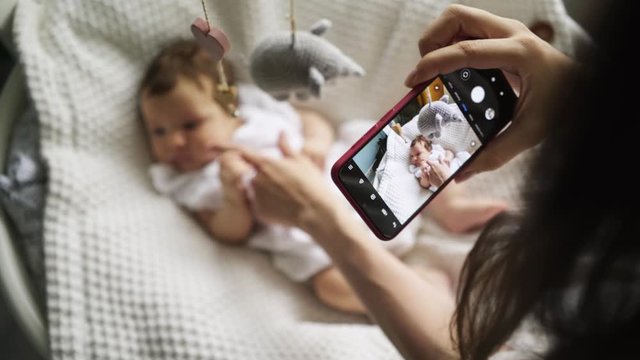 Mother standing near the cradle and taking photo while holding baby's hand. View phone screen, close-up hands with smartphone and child in blur. 