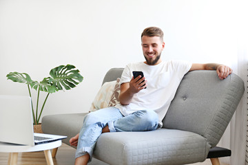 Bearded guy wearing blank white t-shirt & denim pants sitting alone at home on grey textile couch. Young man w/ facial hair in domestic situations. Interior background, copy space, close up, monstera. - Powered by Adobe