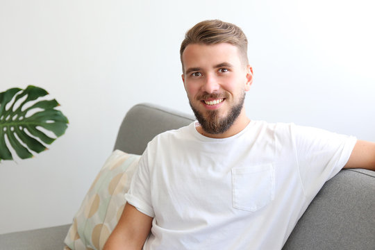 Bearded guy wearing blank white t-shirt & denim pants sitting alone at home on grey textile couch. Young man w/ facial hair in domestic situations. Interior background, copy space, close up, monstera.