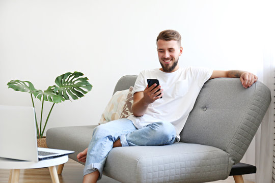 Bearded Guy Wearing Blank White T-shirt & Denim Pants Sitting Alone At Home On Grey Textile Couch. Young Man W/ Facial Hair In Domestic Situations. Interior Background, Copy Space, Close Up, Monstera.