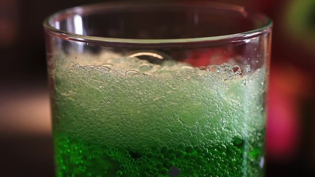 Closeup view how foam disappears in green estragon lemonade in the water glass. Full HD 60fps footage with shallow depth and blurring bokeh.